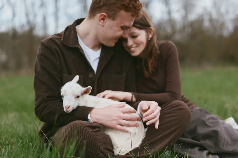 Joyful couple outdoors with a playful goat kid, gentle touch and warm light, relaxed mood, grassy field setting, candid moment, natural colors, simple affection, countryside atmosphere