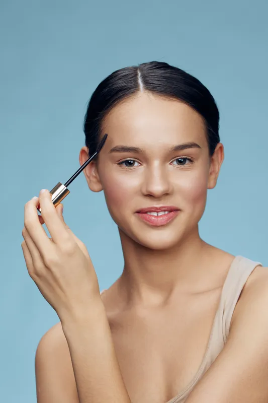 Young woman applying eyebrow gel with a smile against a soft blue background, featuring a natural look and fresh, youthful skin tone