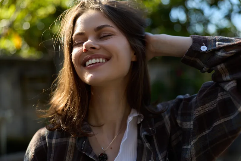 A smiling young woman in a plaid shirt stands outdoors in bright sunlight, capturing a relaxed moment of casual confidence and everyday style with a natural glow