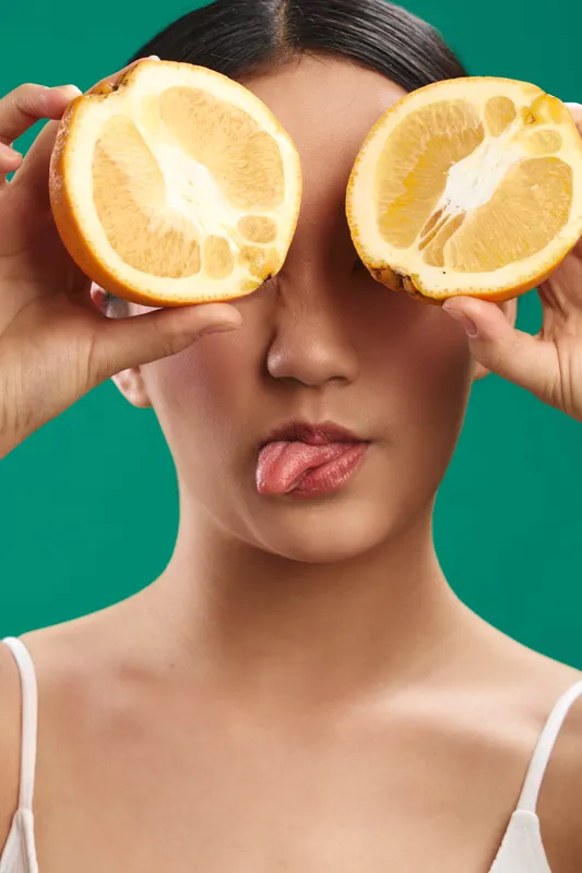 Asian woman playfully holds lemon halves over her eyes, creating a bright and vivid portrait with a fresh, quirky mood. The bold colors and relaxed expression evoke a creative, modern vibe suitable