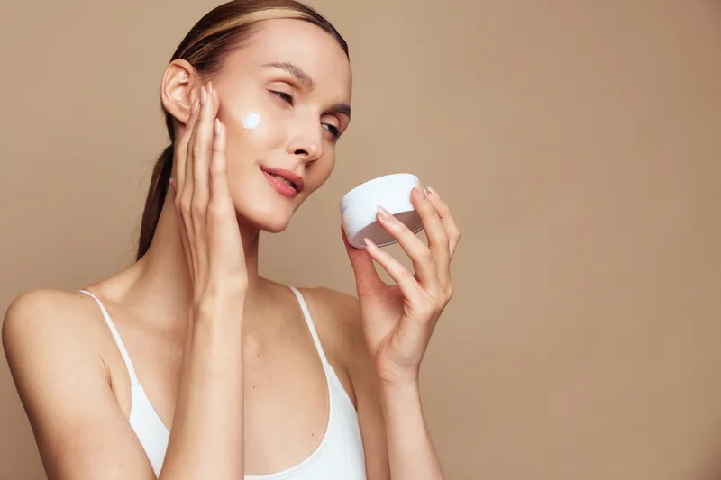 A young woman applies moisturizing cream on her face with her hand while holding a white jar. The neutral background highlights skincare and beauty routine concepts.