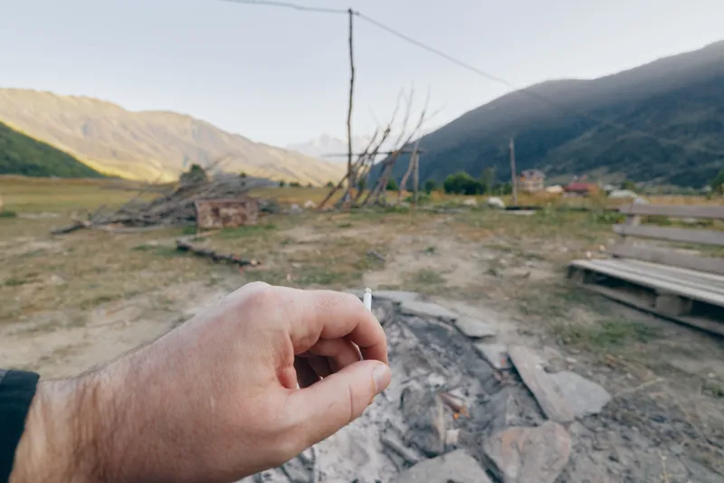 Hand holding cigarette butt above cold campfire ashes in rural mountains near a wooden bench and grassy field, outdoor camping scene with late afternoon light, solitude and quiet nature.