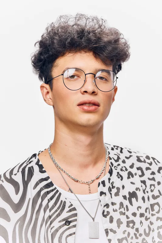 young man with curly hair wearing round glasses and a patterned shirt stands against a white background in a cool modern style portrait
