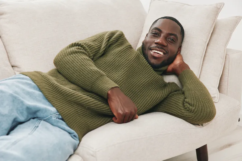 Young Black man in green sweater relaxing on a sofa, smiling joyfully, showcasing comfort and happiness in a cozy indoor setting