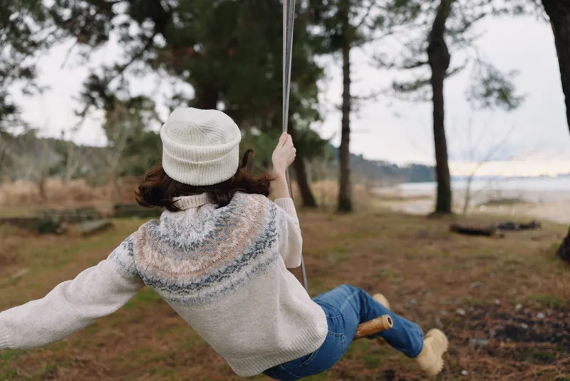 Swinging outdoors among trees by a lake, a person in a sweater and beanie enjoys a peaceful moment of nature, relaxation and leisure in open countryside