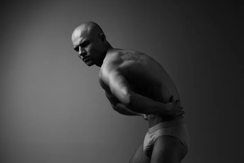 Black and white image of a man in a thoughtful pose, showcasing strong physique and emotional depth, emphasizing contrast and texture in lighting.