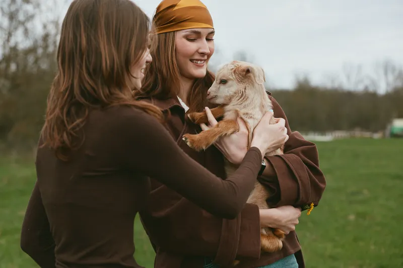 Goat kid moment with woman outdoor lifestyle friendship, two friends cuddle the goat kid, cheerful laughter, sunlit meadow, casual clothing, soft breeze, natural colors, rustic countryside background