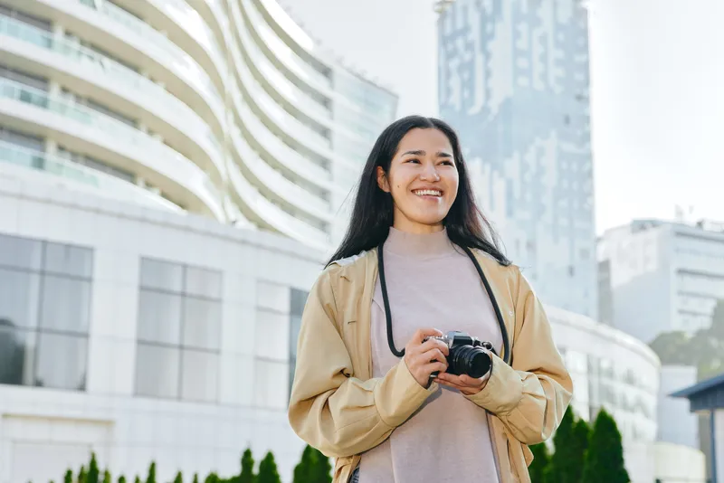 Woman with camera outdoors in urban setting, smiling, posing with lens against modern buildings, lifestyle photography in a vibrant cityscape with daylight and candid charm