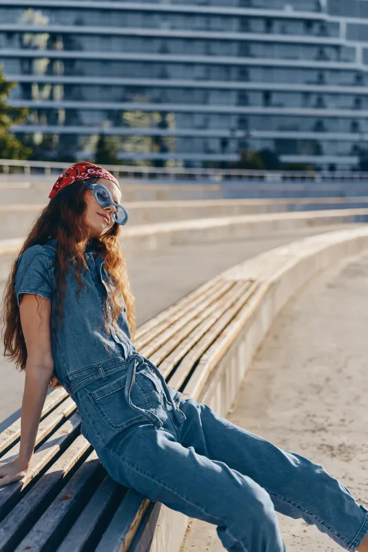 Lifestyle woman in modern boho-western denim street style relaxing on urban bench during sunny day with film color tones, casual and carefree mood captured.