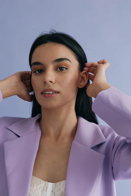 young woman with dark hair wearing lilac blazer poses against soft purple background, showcasing natural makeup and calm expression in modern portrait style