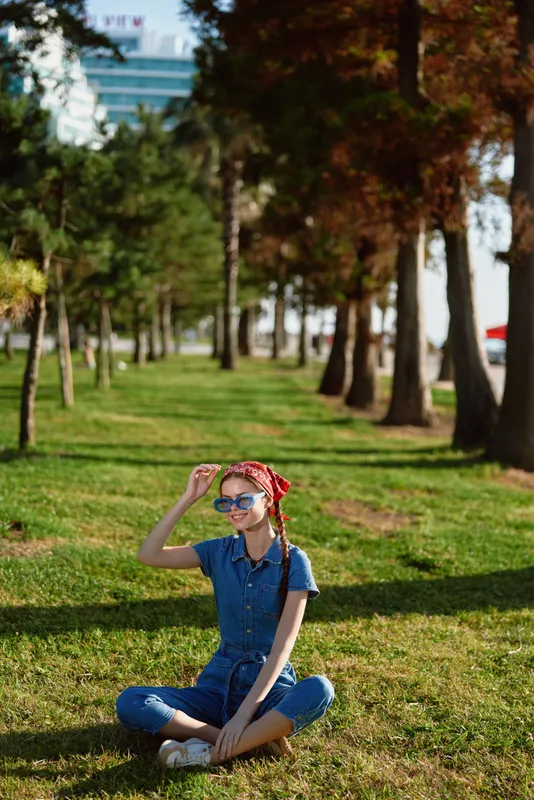 Lifestyle woman in modern boho-western denim street style sitting cross-legged on green grass, wearing blue denim jumpsuit and red headscarf in film color tones.