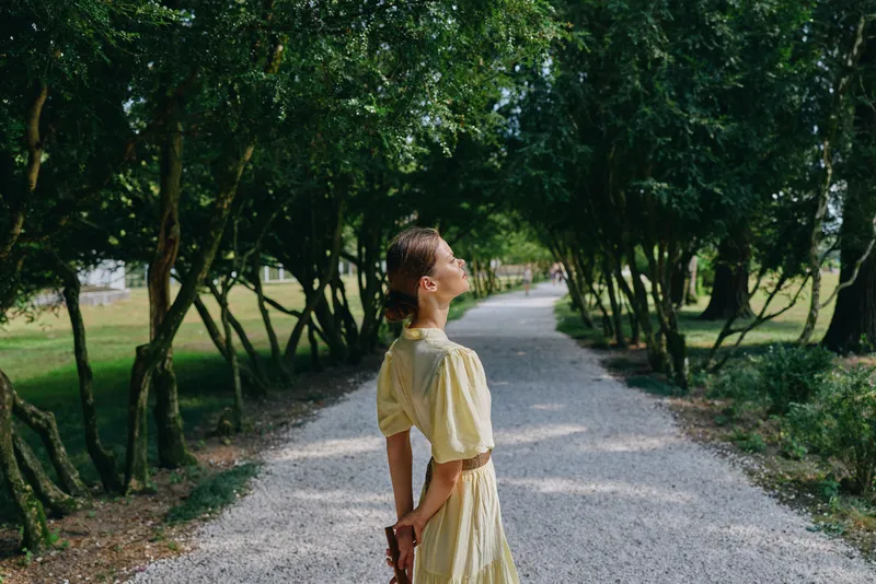 Woman in a yellow dress walks along a gravel path in a sunny park, enjoying a calm moment outdoors, creating a tranquil scene of leisure and nature.