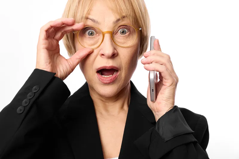 Surprised woman with glasses, holding a smartphone near her ear, business suit, attentive facial expression, communication call, indoor studio lighting, portrait, white background