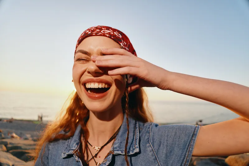 lifestyle woman smiling in modern boho-western denim street style with film color effect, red headscarf, outdoor beach setting, natural light, joyful expression