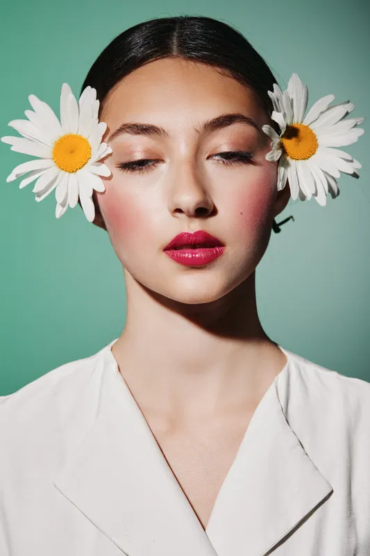 Young woman with flawless skin and vibrant makeup poses with white daisies on both ears. Cotten soft background highlights her fashin coral lipstick and fresh creativity. Beauty concept.