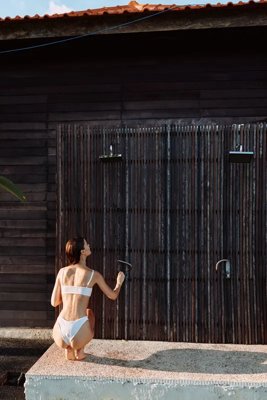 A woman in a white bikini kneels by an outdoor shower, spraying water against a dark wooden wall. A candid lifestyle moment emphasizing relaxation, vacation vibes, and personal care by the water.