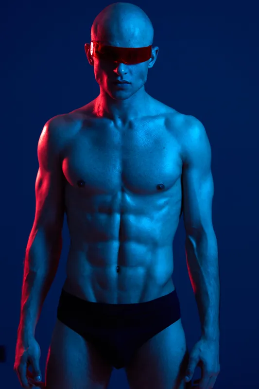 Athletic man posing confidently with sunglasses, showcasing a strong physique against a dramatic blue and red light background, emphasizing modern aesthetics.