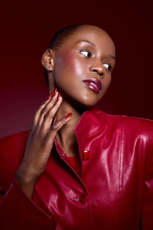 Elegant dark skinned African woman with glossy lips and red nails poses in shiny red leather jacket on red background. Concept of fashion, beauty, and confident female model portrait.
