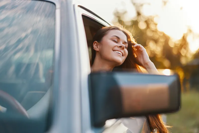 Young woman enjoying a sunny day, smiling from a car window, with golden sunlight and blurred nature background, representing joy and freedom in summer.