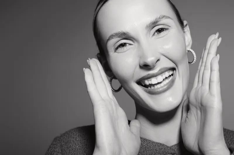 Smiling white woman with light skin touches face with hands. Close-up portrait in monochrome studio lighting.