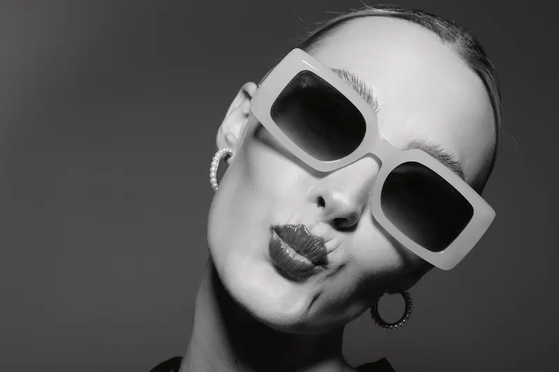 Close-up of a young white woman wearing large sunglasses and hoop earrings making a playful face against a dark background. Concept of fashion and fun.