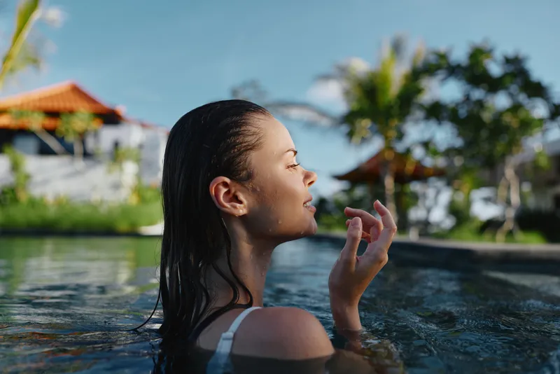 Young woman relaxing in outdoor pool, eyes closed, enjoying peaceful moment, wet hair slicked back, tropical background with palm trees and blue sky, serene atmosphere