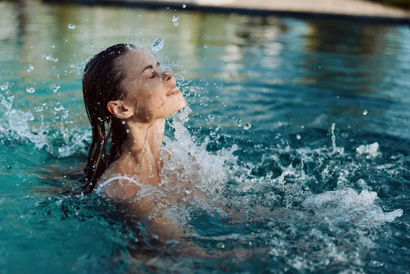 A woman relaxed in a sunlit pool, her face tilted toward the sky as water droplets splash around. A moment of refreshment, calm, and vibrant summer leisure in a casual outdoor setting.