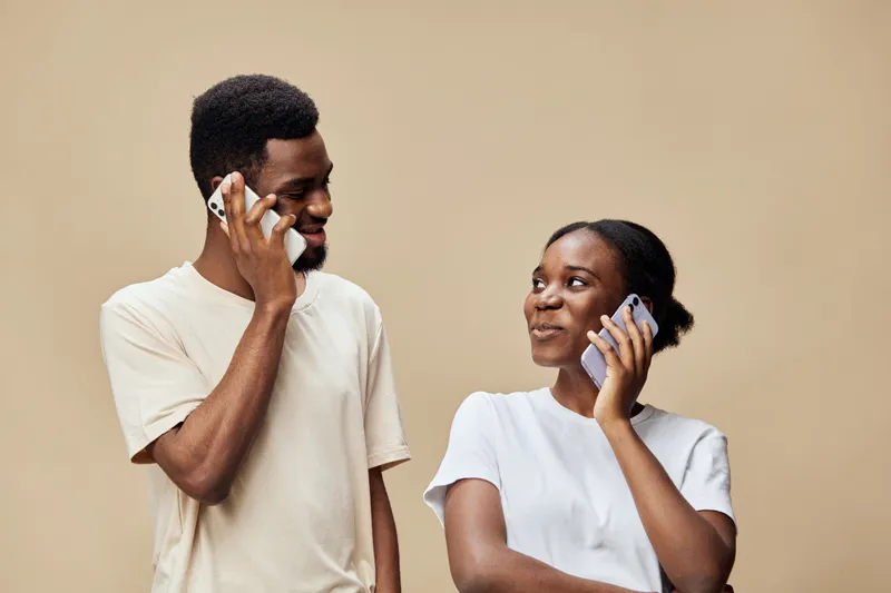 Young African couple communicating joyfully on their smartphones against a neutral beige background, showcasing connection and modern technology