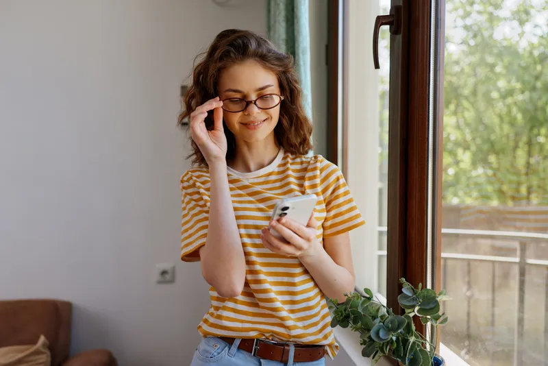 Smiling woman in a striped shirt using a smartphone at home, a casual lifestyle portrait by a sunlit window. Natural light creates a warm, relaxed mood. Great for home wellness moments.