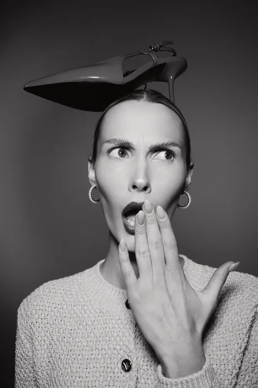 A surprised white woman with pale skin and dark hair holds her hand to her mouth with a high heel shoe balanced on her head in a studio. Concept of fashion and shock.