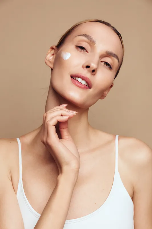 Young woman applies cream on cheek for skincare routine, posing in a beige background, clear skin, calm expression, beauty portrait, lifestyle skincare concept