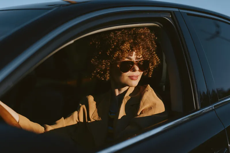 A stylish woman with an afro sits in a car, wearing sunglasses and a tan jacket, exuding retro fashion and confident mood, captured in warm sunlight from outside the window.