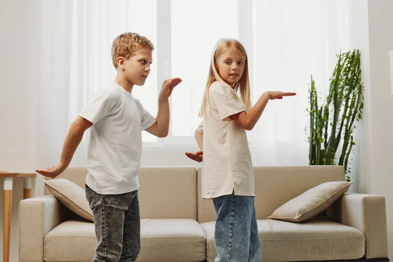 Two children stand back to back playing indoors in casual clothes near sofa and plant in bright living room. Concept of childhood, friendship, playtime, and home activity.
