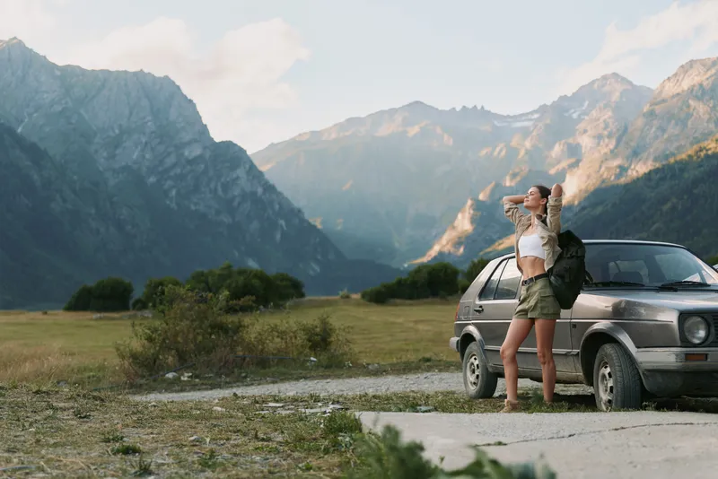 A woman traveler stands beside a rugged car in a mountain landscape, wiping sweat from her brow after a long hike. Adventure and exploration vibe.