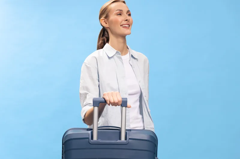 Young woman in casual clothes smiling while holding suitcase handle against blue background. Concept of travel, vacation, and holiday planning with cheerful, relaxed mood.