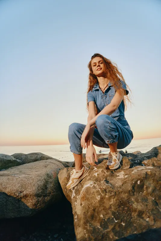 lifestyle woman in modern boho-western denim street style poses confidently on rocks at sunset, capturing a warm film color vibe outdoors by the sea