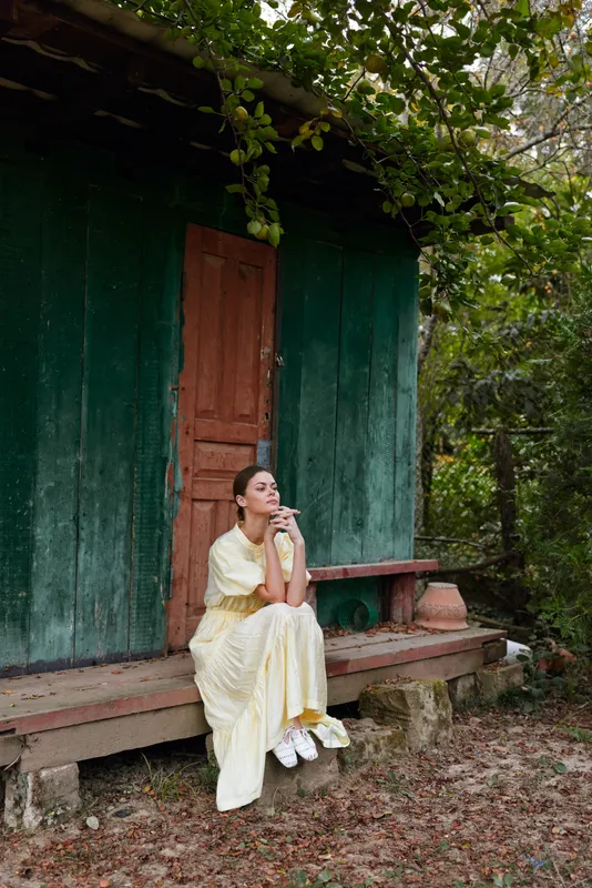 Woman in a yellow dress sits on a wooden veranda outside a green cottage, resting thoughtfully amid leafy surroundings, creating a peaceful outdoor portrait with natural light