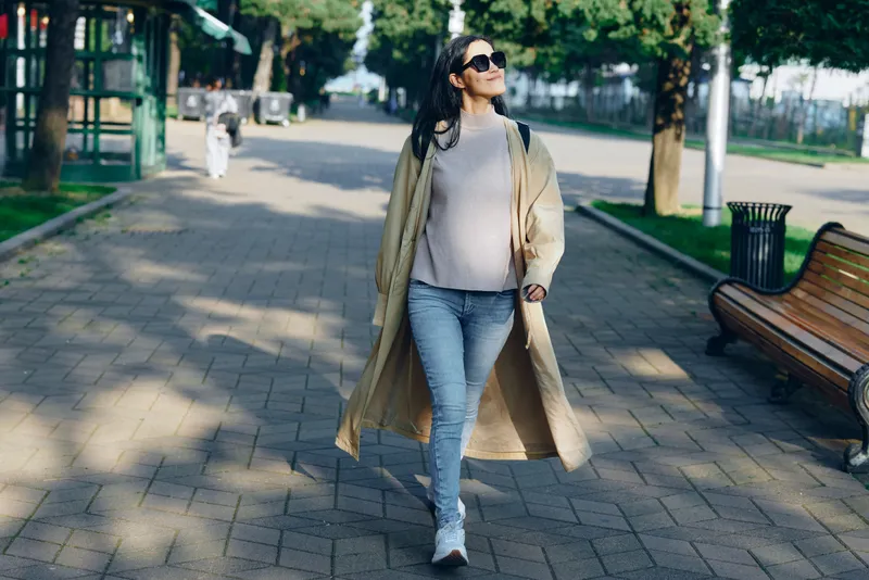 Fashionable woman strolling along a sunlit park path in a long beige coat and jeans, wearing sunglasses, exuding casual confidence during an outdoor urban lifestyle moment