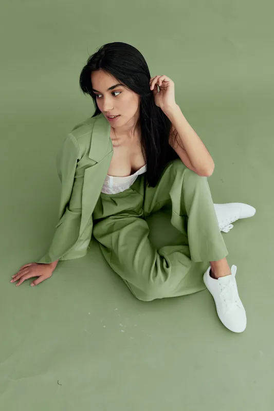 Stylish woman in green pantsuit sitting on green background, wearing white sneakers and lace top, with dark hair and thoughtful expression