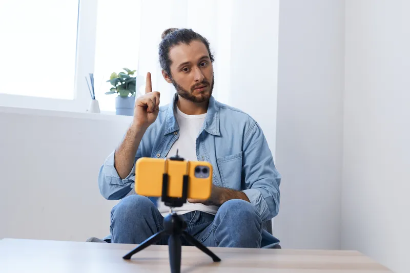 Young man gesturing with a thoughtful expression while recording a video on a smartphone, dressed casually in a denim jacket, indoor bright setting