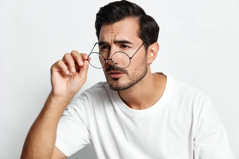 Thoughtful young man adjusting glasses, wearing a plain white t-shirt, looking at the camera with a curious expression against a neutral background. Style concept.