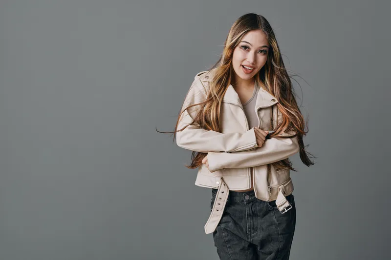 Young woman with long hair wearing beige jacket and black pants standing with arms crossed on gray background. Casual fashion style, confident pose with relaxed expression.