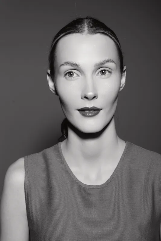 Portrait of a slim white woman with pale skin and dark hair pulled back wearing a sleeveless top on gray background. Studio lighting highlights facial features.