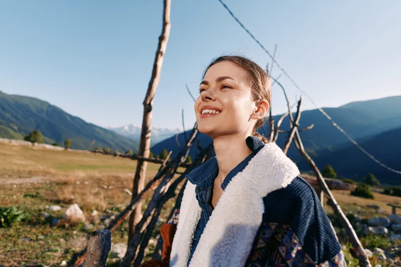 Woman portrait smiling with closed eyes in mountains and nature outdoors, happy traveler enjoying fresh air and sunlight, scenic countryside background and relaxed travel mood.