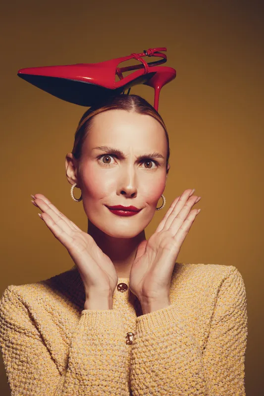 Woman with red high heel shoe on head makes funny face with raised eyebrows and hands near face. Studio portrait on yellow background with fashion and humor concept.