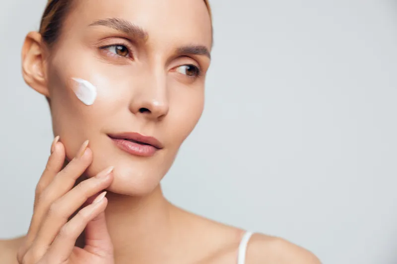 A young woman with clear skin applies white cream on her cheek while gently touching her face. The neutral background emphasizes skincare and beauty routine concepts.
