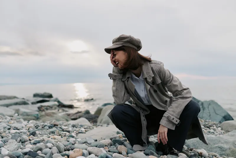 Young woman in stylish coat and hat crouches on rocky beach looking away. Overcast sky and calm sea in background create moody atmosphere and casual outdoor fashion concept.