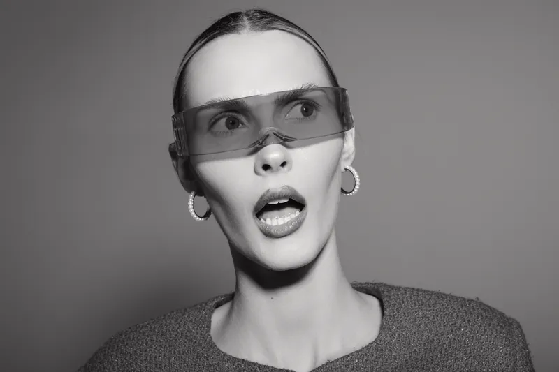 Young Caucasian woman with fair skin wearing transparent visor glasses looks surprised in monochrome studio portrait. Concept for fashion, futuristic, and expression.