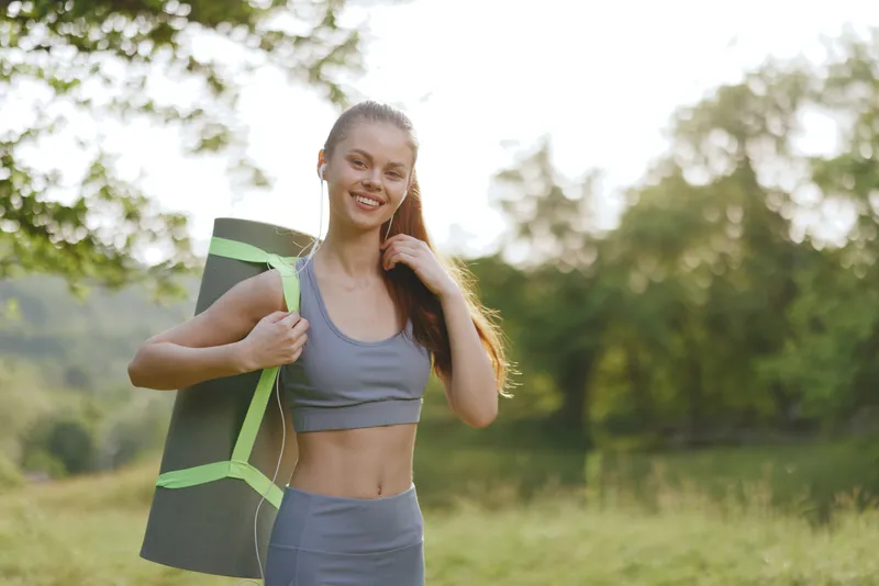 Young female model carrying yoga mat in a serene outdoor setting, wearing a stylish grey workout outfit with a cheerful expression, capturing a healthy lifestyle