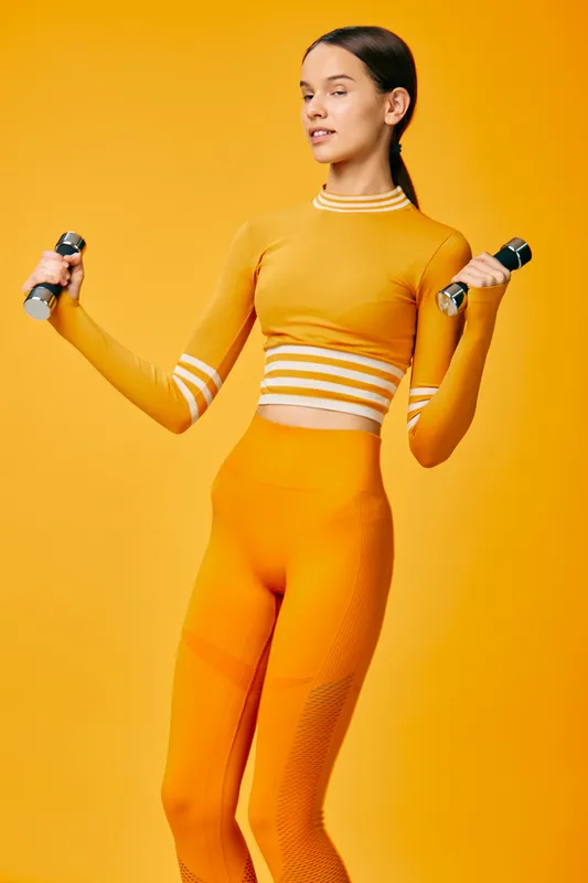 Young fit woman in bright orange matching activewear and leggings lifting silver dumbbells in a monochrome studio background, confident workout pose conveying strength and healthy lifestyle.